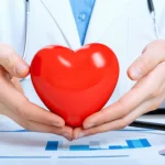 Immagine concettuale che rappresenta la salute cardiovascolare pediatrica: un cuore stilizzato rosso brillante protetto da mani di bambino e adulto sovrapposte, su uno sfondo sfocato di grafici medici e strumenti diagnostici. Lente macro 90mm, illuminazione da studio controllata per enfatizzare il cuore, alta definizione dei dettagli, colori caldi e rassicuranti.