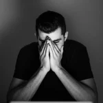 Ritratto di uno studente universitario visibilmente preoccupato, con la testa tra le mani, seduto davanti a un laptop in una stanza poco illuminata, simbolo dell'isolamento e dello stress da quarantena COVID-19. Obiettivo 35mm, bianco e nero con grana fine, stile film noir per accentuare il dramma e l'introspezione.