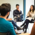 Un'aula di un Recovery College luminosa e accogliente, con persone di età diverse sedute in cerchio che discutono animatamente. Alcuni prendono appunti, altri gesticolano. L'atmosfera è collaborativa e positiva. Obiettivo prime da 35mm, luce naturale che entra da grandi finestre, colori vivaci ma non saturi, profondità di campo media.