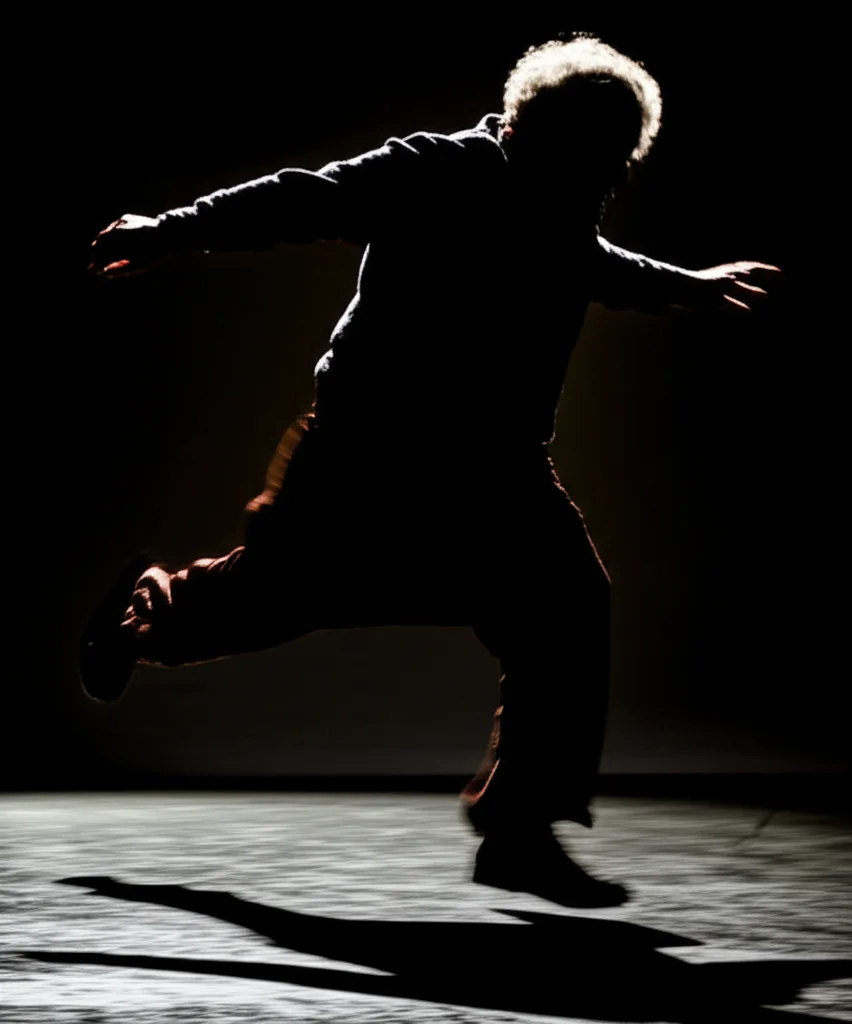 Immagine dinamica che mostra la silhouette di una persona anziana che perde l'equilibrio su una superficie scivolosa, con le braccia che si muovono rapidamente per tentare di recuperare la stabilità. L'immagine è catturata con un teleobiettivo zoom 200mm, fast shutter speed, per congelare l'azione, con un leggero motion blur sullo sfondo per enfatizzare il movimento. Illuminazione drammatica laterale.