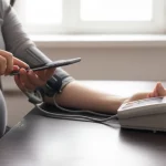 Primo piano di una donna incinta sorridente che usa uno smartphone per controllare i dati della sua pressione sanguigna tramite un'app dedicata, accanto a un misuratore di pressione digitale domestico appoggiato su un tavolo. Luce naturale morbida proveniente da una finestra laterale, profondità di campo ridotta che sfoca lo sfondo, obiettivo 50mm prime lens.