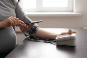 Primo piano di una donna incinta sorridente che usa uno smartphone per controllare i dati della sua pressione sanguigna tramite un'app dedicata, accanto a un misuratore di pressione digitale domestico appoggiato su un tavolo. Luce naturale morbida proveniente da una finestra laterale, profondità di campo ridotta che sfoca lo sfondo, obiettivo 50mm prime lens.