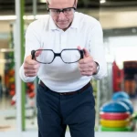Un anziano sorridente ma concentrato prova degli occhiali stroboscopici futuristici, in piedi con sicurezza su una pedana di equilibrio. Sfondo leggermente sfocato di un ambiente moderno e luminoso tipo palestra riabilitativa. Ritratto con obiettivo 35mm, profondità di campo, toni caldi e rassicuranti, luce naturale morbida.