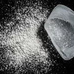 Immagine macro ad alta definizione di polvere fine di membrana amniotica bovina liofilizzata sparsa su una superficie sterile scura, accanto a un frammento traslucido e umido della stessa membrana fresca. Illuminazione da studio controllata che enfatizza la differenza di texture e colore tra le due forme, obiettivo macro 90mm, focus selettivo sulla polvere.
