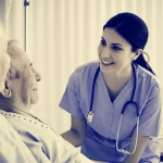 Studentessa infermiera sorridente e compassionevole interagisce con un paziente in un ambiente ospedaliero luminoso. Fotografia ritratto, obiettivo da 35mm, toni seppia e blu duotone, profondità di campo che evidenzia i soggetti.