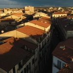 Veduta aerea di un vecchio quartiere urbano con tetti antichi e strade strette, parzialmente in ombra e parzialmente illuminato dal sole, obiettivo grandangolare 20mm, per trasmettere un senso di vulnerabilità e potenziale, con un focus nitido sulle texture degli edifici storici e un cielo drammatico al tramonto.