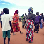 Fotografia di persone in un contesto rurale dell'Africa Orientale, magari vicino a un centro sanitario mobile o durante una campagna di sensibilizzazione. Uomini e donne di età diverse. Stile reportage, obiettivo 35mm, profondità di campo che isola un piccolo gruppo ma lascia intravedere la comunità, luce naturale filtrata, colori caldi e terrosi con un tocco di blu e verde per un effetto duotone sottile. L'immagine deve trasmettere sia la vulnerabilità che la resilienza delle popolazioni mobili.