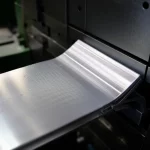 Immagine fotorealistica di un componente industriale in acciaio piegato a U (profilato per sedile auto) appena uscito da una pressa industriale, con evidenza della precisione dell'angolo ottenuto grazie a sistemi di controllo avanzati. Obiettivo prime 35mm, profondità di campo ridotta per isolare il pezzo, illuminazione industriale drammatica che esalta le superfici metalliche.