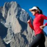 Atleta donna allenata (runner) su un sentiero di montagna a circa 4000 metri, luce naturale intensa del primo pomeriggio, teleobiettivo 200mm con fast shutter speed per catturare il movimento, focus nitido sull'atleta, sfondo montuoso imponente leggermente sfocato per dare profondità, cielo azzurro intenso tipico dell'alta quota.