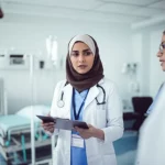 A diverse group of emergency nurses in an Iranian hospital, engaged in a collaborative huddle. One nurse is speaking while others listen attentively, some nodding. The background shows a busy but organized emergency room. The image should convey professionalism, teamwork, and a sense of calm amidst potential chaos. Portrait photography style, 35mm lens, with a subtle duotone of blue and grey to enhance a serious yet hopeful mood. Depth of field is used to keep the nurses in sharp focus.