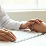 Fotografia realistica, scattata con obiettivo prime 50mm, che mostra le mani di un medico (con camice bianco) e di un paziente (con abbigliamento casual) appoggiate su un modulo di consenso informato aperto su un tavolo in un ambiente ospedaliero luminoso e rassicurante. Profondità di campo che sfoca leggermente lo sfondo (una finestra o una parete chiara), mettendo in risalto l'interazione focalizzata sul documento. Tonalità neutre e calde.