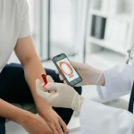 Fotografia realistica, un medico con camice bianco esamina attentamente un neo sulla pelle di un paziente utilizzando uno smartphone con un'app per la diagnosi del cancro della pelle. L'ambiente è un ambulatorio medico ben illuminato. Obiettivo prime 50mm, profondità di campo, illuminazione professionale da studio.
