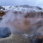 landscape wide angle 10mm, sharp focus, a dramatic view of a high-elevation volcanic fumarole field with steam rising against a backdrop of snow-capped peaks, conveying extreme conditions