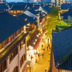 Veduta aerea di un quartiere Shikumen di Shanghai al crepuscolo, che mostra un mix di tetti tradizionali in mattoni grigi e spazi verdi rigenerati, con luci calde che illuminano i vicoli e persone che passeggiano. Obiettivo grandangolare 20mm, lunga esposizione per cieli suggestivi e scie luminose, focus nitido sull'architettura e sull'atmosfera vibrante.