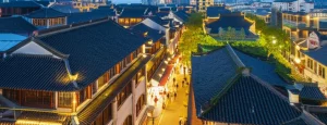 Veduta aerea di un quartiere Shikumen di Shanghai al crepuscolo, che mostra un mix di tetti tradizionali in mattoni grigi e spazi verdi rigenerati, con luci calde che illuminano i vicoli e persone che passeggiano. Obiettivo grandangolare 20mm, lunga esposizione per cieli suggestivi e scie luminose, focus nitido sull'architettura e sull'atmosfera vibrante.