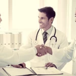 Fotografia realistica di un medico di base sorridente e un'infermiera domiciliare che si stringono la mano sopra una cartella clinica aperta su una scrivania in un ambulatorio luminoso. Simboleggia collaborazione e intesa. Obiettivo prime 50mm, luce naturale morbida da una finestra laterale, colori caldi e neutri, duotone seppia e grigio chiaro per un tocco professionale ma umano.