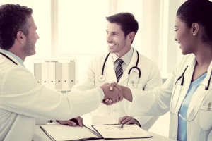 Fotografia realistica di un medico di base sorridente e un'infermiera domiciliare che si stringono la mano sopra una cartella clinica aperta su una scrivania in un ambulatorio luminoso. Simboleggia collaborazione e intesa. Obiettivo prime 50mm, luce naturale morbida da una finestra laterale, colori caldi e neutri, duotone seppia e grigio chiaro per un tocco professionale ma umano.