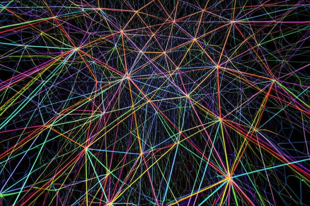 Illustrazione concettuale avanzata di una simulazione di trasporto di particelle su una mesh poliedrica Voronoi tridimensionale. Linee di flusso luminose e colorate attraversano le celle trasparenti della mesh, suggerendo movimento e direzione. Wide-angle lens, 10mm, long exposure effect per creare scie luminose fluide, sharp focus sulla struttura della mesh Voronoi, sfondo scuro per enfatizzare il flusso.