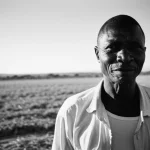 Ritratto intenso di un agricoltore anziano del Ghana settentrionale che guarda verso l'orizzonte tra i suoi campi, luce calda del tardo pomeriggio, obiettivo 50mm, profondità di campo ridotta per sfocare lo sfondo, bianco e nero ad alto contrasto, stile film noir.