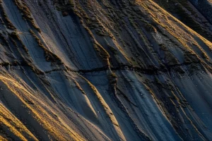 Fotografia paesaggistica, obiettivo grandangolare 20mm, un pendio roccioso stratificato con un evidente interstrato più scuro e morbido visibile a mezzacosta. Luce drammatica del tardo pomeriggio che evidenzia le texture e le stratificazioni. Messa a fuoco nitida sull'intera scena, mostrando la potenziale instabilità legata all'interstrato.