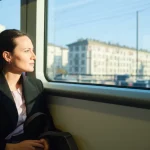 Una persona sorridente che viaggia comodamente su un treno moderno e poco affollato, guardando fuori dal finestrino un paesaggio urbano soleggiato. Obiettivo prime 35mm, profondità di campo per mettere a fuoco la persona e sfocare leggermente lo sfondo, luce naturale che entra dal finestrino.