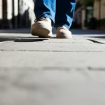 Immagine concettuale fotorealistica: vista soggettiva dei piedi di una persona anziana che cammina su un marciapiede, la parte inferiore dell'immagine è leggermente sfocata o oscurata per rappresentare la perdita del campo visivo inferiore. Obiettivo grandangolare, 24mm, messa a fuoco sui piedi e sul terreno immediatamente davanti, luce diurna naturale.