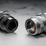 Immagine fotorealistica di un confronto tra un terminale (lug) in CFRP nero lucido e uno in alluminio grigio opaco, posizionati su un banco da laboratorio metallico. Luce da studio laterale che evidenzia le texture dei materiali. Obiettivo prime 35mm, profondità di campo ridotta per mettere a fuoco i dettagli dei lug.