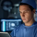 A close-up portrait of a male surgeon in scrubs looking intently at a tablet displaying medical data, 35mm portrait, depth of field, controlled lighting.