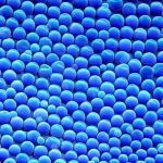 Primo piano fotorealistico di nanoparticelle core-shell-shell che emettono una brillante luce blu-viola, sospese in una soluzione limpida. L'immagine cattura la luminescenza intensa, con dettagli nitidi delle particelle quasi individuali. Obiettivo macro 100mm, illuminazione laterale controllata per esaltare la brillantezza, alta definizione.