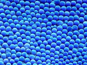 Primo piano fotorealistico di nanoparticelle core-shell-shell che emettono una brillante luce blu-viola, sospese in una soluzione limpida. L'immagine cattura la luminescenza intensa, con dettagli nitidi delle particelle quasi individuali. Obiettivo macro 100mm, illuminazione laterale controllata per esaltare la brillantezza, alta definizione.