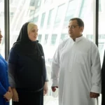 Un gruppo diversificato di adulti a Makkah, Arabia Saudita, in un contesto urbano moderno che suggerisce un centro sanitario o comunitario, alcuni visibilmente sovrappeso, impegnati in una conversazione sulla salute e il benessere. Fotografia di ritratto di gruppo, obiettivo prime 35mm, luce naturale morbida proveniente da finestre, profondità di campo media per mantenere a fuoco le persone ma sfocare leggermente lo sfondo architettonico, atmosfera seria ma speranzosa.
