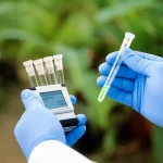 Immagine concettuale di diagnostica rapida in contesto a risorse limitate: mani guantate di un operatore sanitario che tengono un piccolo dispositivo diagnostico portatile (simile a un fluorimetro RPA) con provette luminose, sfondo sfocato di una clinica rurale o paesaggio tropicale. Obiettivo prime 50mm, profondità di campo ridotta per focalizzare sul dispositivo e le mani.