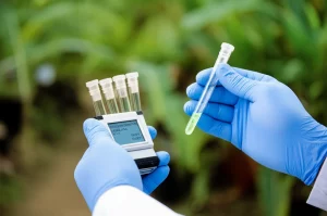 Immagine concettuale di diagnostica rapida in contesto a risorse limitate: mani guantate di un operatore sanitario che tengono un piccolo dispositivo diagnostico portatile (simile a un fluorimetro RPA) con provette luminose, sfondo sfocato di una clinica rurale o paesaggio tropicale. Obiettivo prime 50mm, profondità di campo ridotta per focalizzare sul dispositivo e le mani.