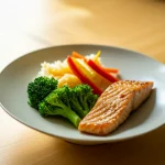 Un piatto di cibo giapponese semplice ma nutriente (riso bianco in una ciotola, verdure saltate colorate, un pezzo di pesce grigliato) disposto su una tavola di legno in un ambiente luminoso che suggerisce una casa temporanea ma accogliente. Fotografia still life, lente macro 85mm, luce naturale morbida laterale, alta definizione, focus preciso sul cibo, profondità di campo ridotta per sfocare leggermente lo sfondo.