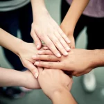 Immagine fotorealistica di un gruppo eterogeneo di persone che uniscono le mani al centro, simboleggiando la forza delle coalizioni e della collaborazione per la giustizia sociale. Scattata con un obiettivo da 35mm, con una profondità di campo che sfoca leggermente lo sfondo per concentrarsi sulle mani unite, utilizzando una palette di colori caldi e accoglienti.