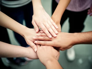 Immagine fotorealistica di un gruppo eterogeneo di persone che uniscono le mani al centro, simboleggiando la forza delle coalizioni e della collaborazione per la giustizia sociale. Scattata con un obiettivo da 35mm, con una profondità di campo che sfoca leggermente lo sfondo per concentrarsi sulle mani unite, utilizzando una palette di colori caldi e accoglienti.
