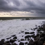 Immagine fotorealistica, scattata con obiettivo grandangolare da 18mm, di una costa indiana drammaticamente illuminata durante un evento meteorologico estremo combinato, con onde imponenti che si infrangono e nuvole temporalesche cariche di pioggia all'orizzonte, lunga esposizione per enfatizzare il movimento dell'acqua e delle nuvole.