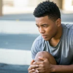 Young male athlete looking thoughtful or reflective about a knee injury, 35mm portrait, depth of field, natural light.