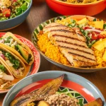Un tavolo vivace apparecchiato con una varietà di piatti colorati rappresentativi delle diverse cucine ispaniche/latine (tacos, riso e fagioli, platano fritto, insalata di quinoa, pesce alla griglia). Luce calda e controllata che mette in risalto le texture dei cibi, obiettivo macro 85mm, alta definizione, focus selettivo su un piatto centrale.