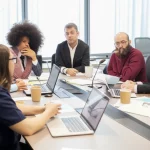 Un gruppo eterogeneo di docenti universitari, uomini e donne di età diverse, seduti attorno a un tavolo moderno in un'aula luminosa. Stanno discutendo animatamente ma in modo costruttivo, con appunti, laptop e prototipi semplici sparsi sul tavolo, rappresentando una sessione di Design Based Learning Study (DBLS). L'atmosfera è collaborativa e focalizzata sull'apprendimento collettivo. Fotografia di ritratti di gruppo, obiettivo prime 35mm, profondità di campo media per mantenere a fuoco il gruppo ma sfocare leggermente lo sfondo dell'aula universitaria, illuminazione naturale morbida.