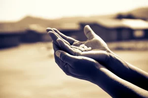 Immagine fotorealistica di forte impatto emotivo: una mano tesa che offre una compressa antiretrovirale a un'altra mano in un contesto rurale dell'Africa subsahariana, simboleggiando la speranza e la sfida dell'accesso alle cure. Obiettivo prime 35mm, luce soffusa e calda, profondità di campo che mette a fuoco le mani e la compressa, con lo sfondo leggermente sfocato che suggerisce un villaggio. Toni seppia e blu duotone per un effetto intenso ma speranzoso.