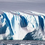 Immagine fotorealistica di una sezione trasversale di una piattaforma di ghiaccio antartica nella zona di ancoraggio. Si vedono chiaramente gli strati di ghiaccio che si inclinano progressivamente con la profondità, passando da orizzontali a molto inclinati. Obiettivo per paesaggi, 20mm, luce diffusa per dettaglio uniforme, messa a fuoco nitida su tutta la sezione.