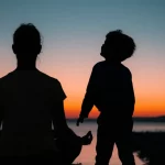 Silhouette di una persona adulta in meditazione al tramonto con sovrapposta l'immagine sbiadita di un bambino che guarda verso l'alto, fotografia paesaggistica, obiettivo grandangolare 24mm, lunga esposizione per nuvole soffuse.