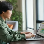 Immagine fotorealistica di un caregiver vietnamita di mezza età che utilizza un laptop seduto a un tavolo in una stanza luminosa. Sullo schermo del laptop è visibile l'interfaccia di un programma educativo web. Il caregiver ha un'espressione concentrata ma calma. Obiettivo prime 50mm, luce naturale morbida proveniente da una finestra laterale, profondità di campo media che mantiene a fuoco sia il caregiver che lo schermo, colori naturali e caldi.
