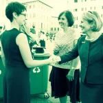 Fotografia realistica di una donna sindaco sorridente che stringe la mano a cittadini in una piazza italiana pulita e verde, con bidoni della raccolta differenziata ben visibili. Obiettivo da 35mm, luce naturale, effetto duotone seppia e verde acqua per un tocco vintage ma moderno.