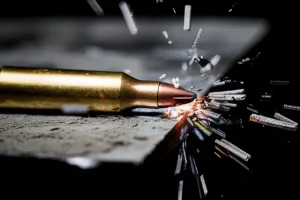Primo piano di un proiettile ad alta velocità che sta per impattare una spessa lastra di metallo, con scintille e frammenti che iniziano a staccarsi dalla superficie al punto di impatto. Obiettivo teleobiettivo zoom 200mm, alta velocità dell'otturatore per congelare l'azione, tracciamento del movimento, illuminazione drammatica laterale per esaltare texture e deformazione, bianco e nero con un singolo colore d'accento per le scintille.