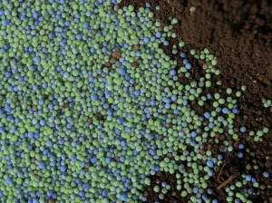 Primo piano fotorealistico di granuli di PET riciclato colorato (trasparente, verde, blu) mescolati con terreno argilloso scuro e umido, simboleggiando l'integrazione dei rifiuti nella stabilizzazione del suolo. Illuminazione laterale drammatica che crea ombre e mette in risalto le texture. Obiettivo macro 100mm, alta definizione dei dettagli, profondità di campo ridotta.