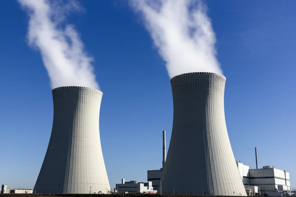 Una centrale nucleare moderna e pulita vista dall'esterno, con torri di raffreddamento che emettono vapore bianco contro un cielo azzurro sereno, fotografia grandangolare, 10mm, lunga esposizione per nuvole setose, messa a fuoco nitida, che simboleggia l'energia e la ricerca scientifica.