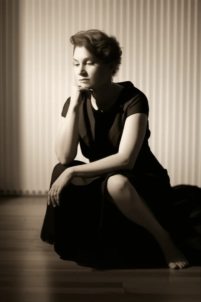 Un ritratto di una persona di mezza età, seduta in una stanza scarsamente illuminata, con un'espressione pensierosa ma con un accenno di speranza. L'illuminazione dovrebbe creare un effetto chiaroscuro, quasi film noir, per sottolineare la lotta interiore. Obiettivo da 35mm, profondità di campo per mettere a fuoco il soggetto e sfocare leggermente lo sfondo. Toni seppia e grigio per un'atmosfera introspettiva.