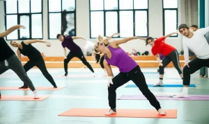 Gruppo eterogeneo di adolescenti con diabete tipo 1 (alcuni con sensori glicemici visibili sul braccio) che ridono e parlano mentre fanno stretching leggero su tappetini colorati in una palestra luminosa. Fotografia di gruppo, prime lens 35mm, profondità di campo media per includere il gruppo ma con sfondo leggermente sfocato, luce naturale dalle finestre.